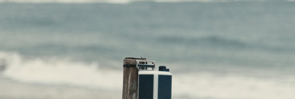 An Obscape monitoring station on a post on a sandy beach, with the coastline and a city skyline behind