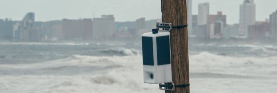An Obscape coastal monitoring station mounted on a pole, overlooking a stormy sea with a city skyline behind