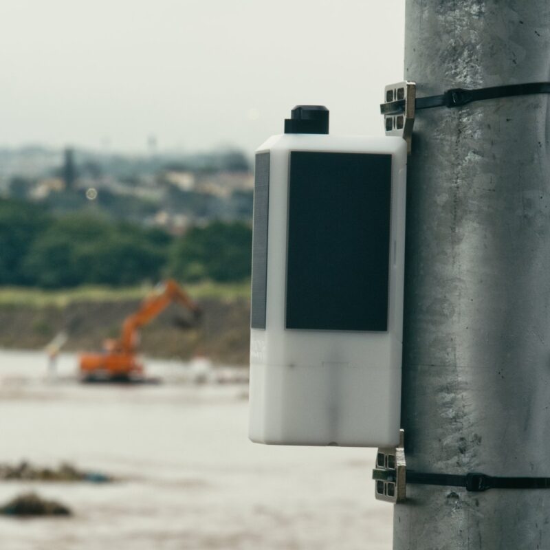 An Obscape water-level monitoring station mounted on a pole above a river, with earthmoving equipment in the background