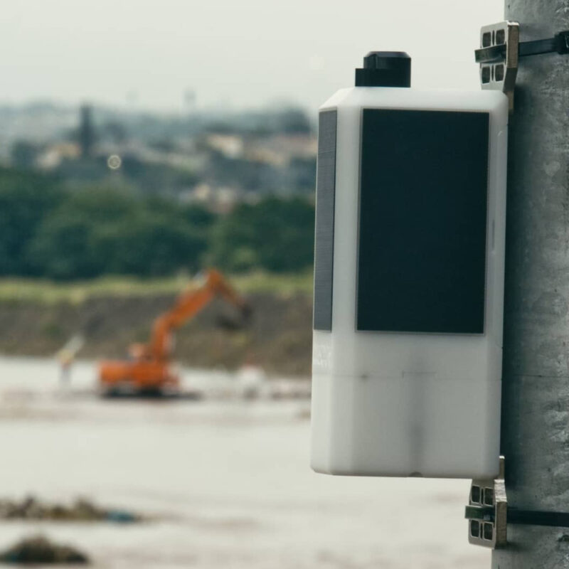 An Obscape water-level monitoring station on a pole above a swollen, sediment-laden river, with an excavator working on the bank behind it